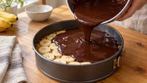 Dunkle Schokoladen-Ganache wird aus einer Schüssel über Bananenscheiben in einer Springform gegossen.